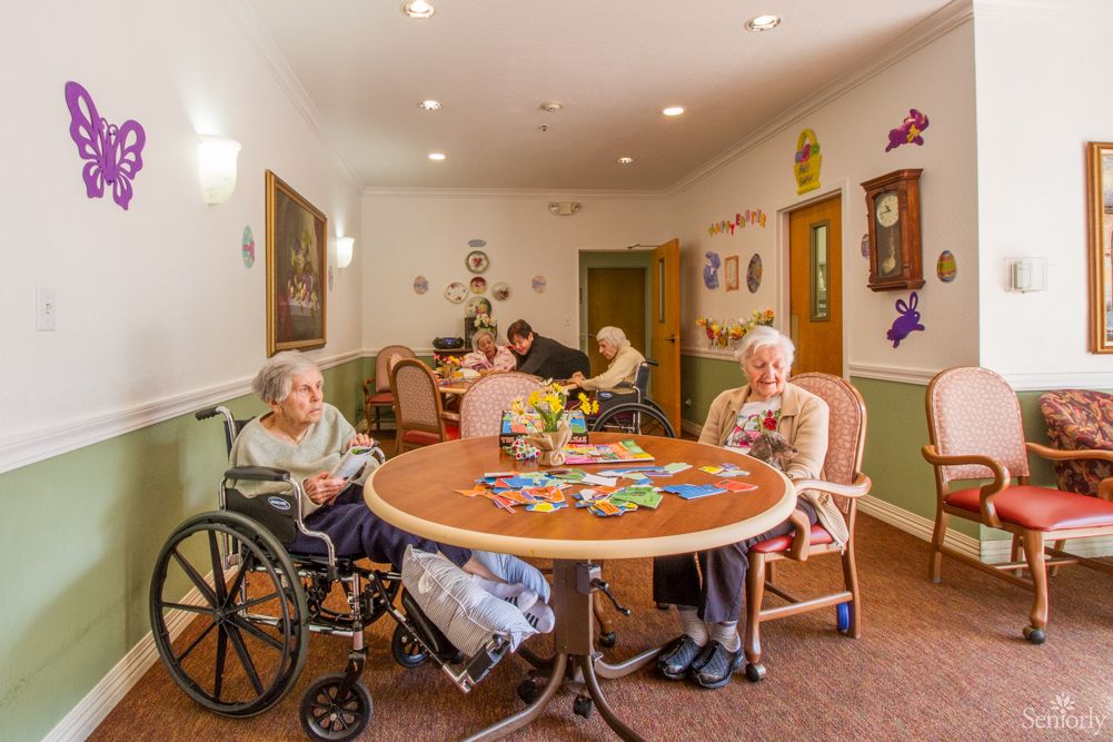 Elderly residents gathered around a table in a nursing home community room.