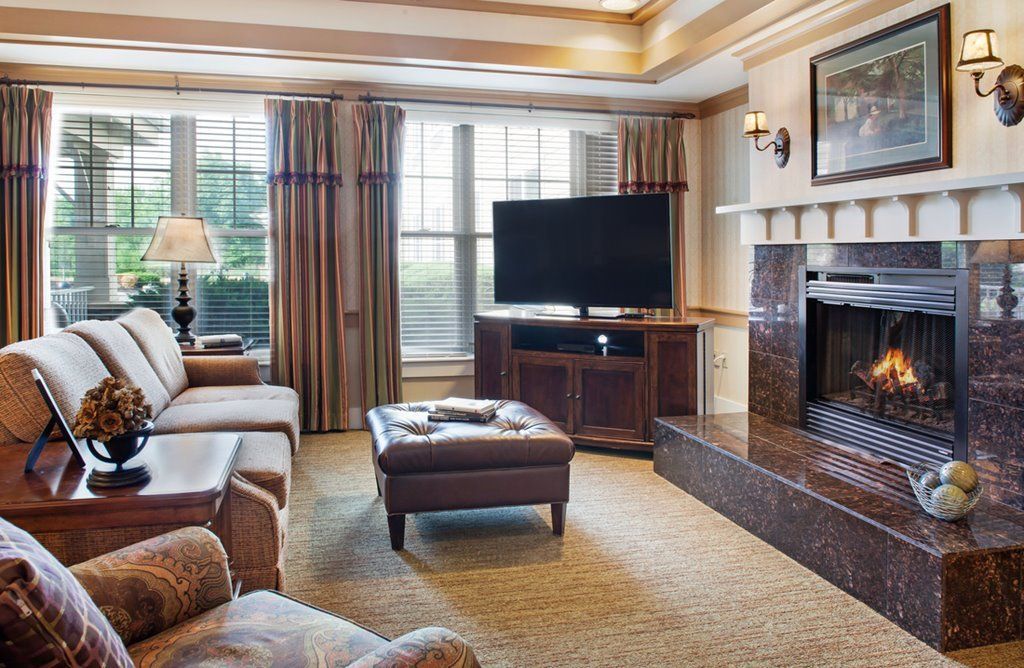 A cozy living room with a television and fireplace, featuring comfortable seating and large windows.