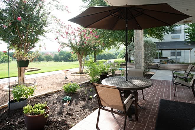 Outdoor patio area with a table, chairs, and an umbrella in a garden setting on a sunny day.