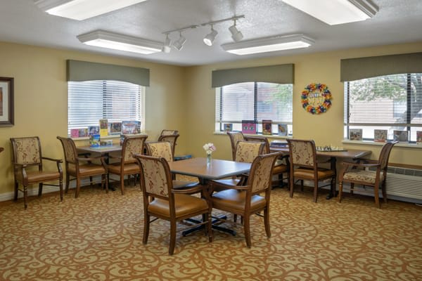 A well-lit dining room with tables and chairs arranged neatly, featuring large windows and warm decor.