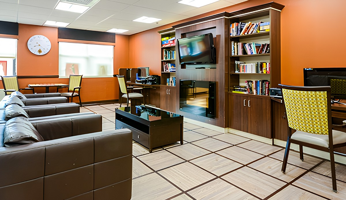 A modern office interior featuring comfortable seating, bookshelves, computer desks, and a wall-mounted television.