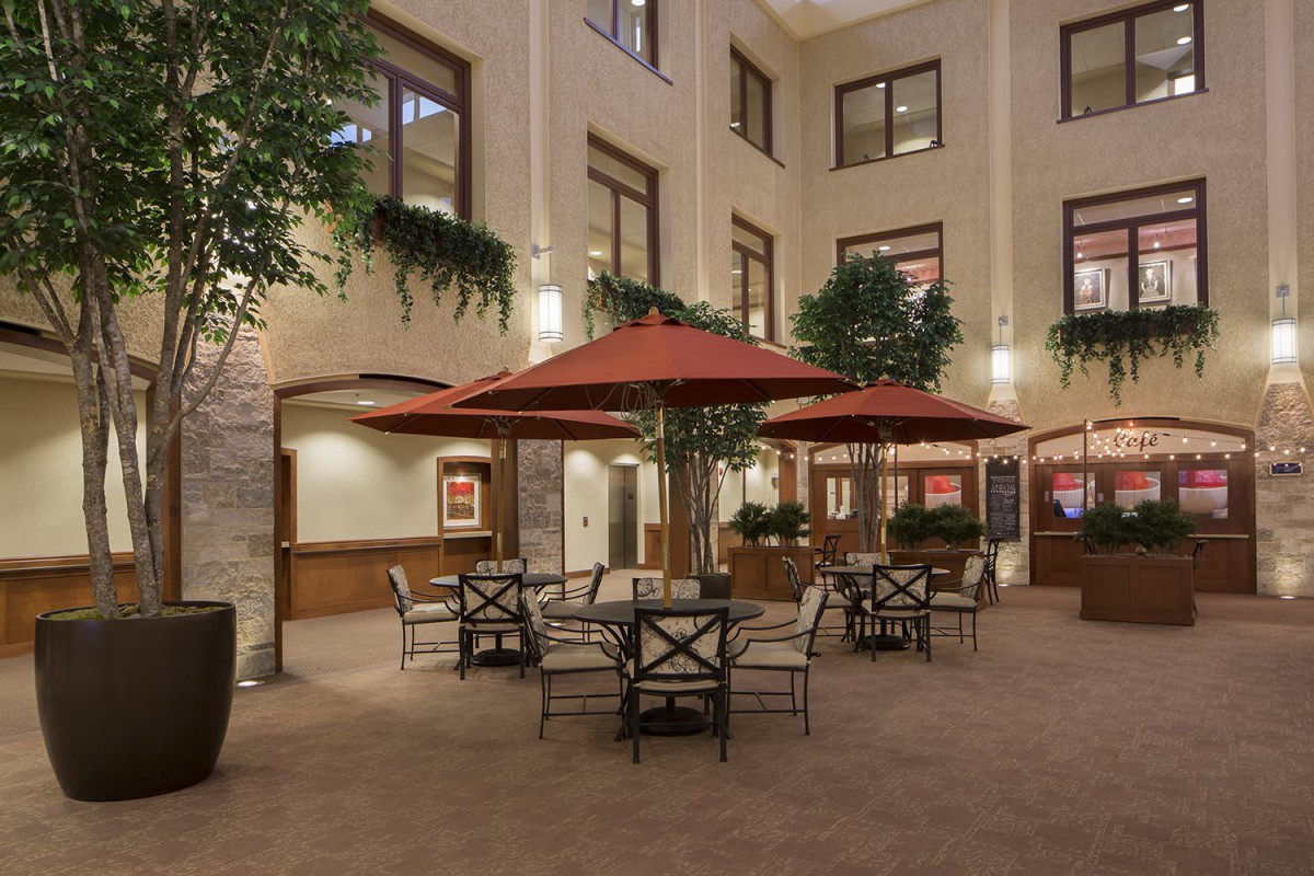 An indoor courtyard with tables, chairs, and red umbrellas surrounded by greenery.