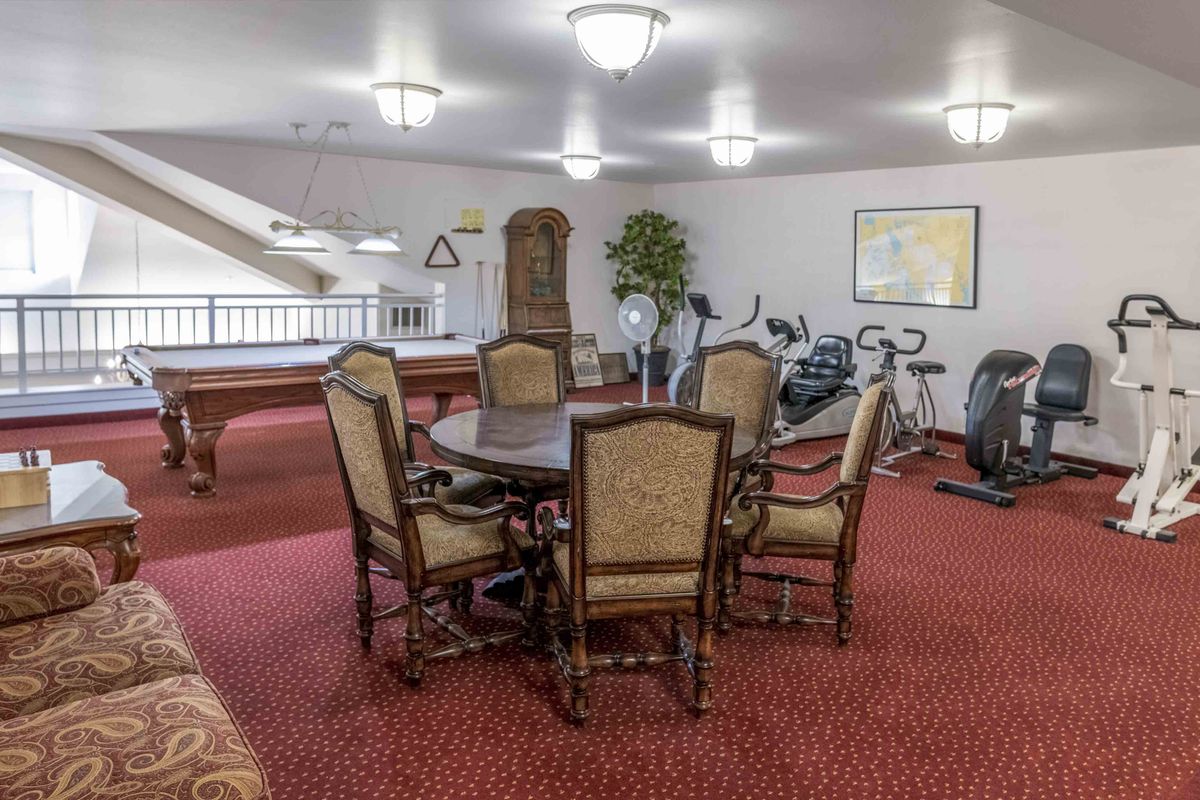 A multipurpose room featuring a round wooden table with chairs, exercise machines, and a billiards table on red carpeted flooring.