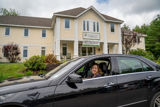 A smiling individual seated in a black car outside a charming building.