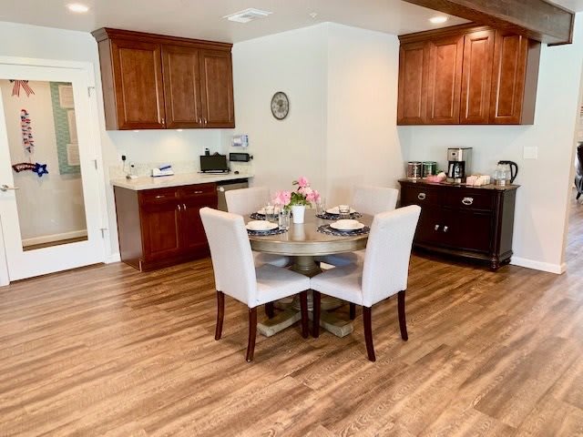 A modern kitchen and dining area with wooden floors, featuring a round table set for four, surrounded by white upholstered chairs. The space includes dark wood cabinets and a coffee station.