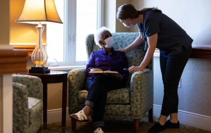 A caregiver attending to an elderly person sitting in a chair, both wearing masks.