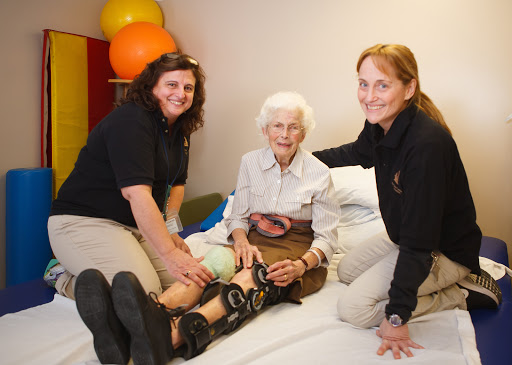 A caregiver assisting an elderly woman with therapy exercises, supported by a second caregiver.