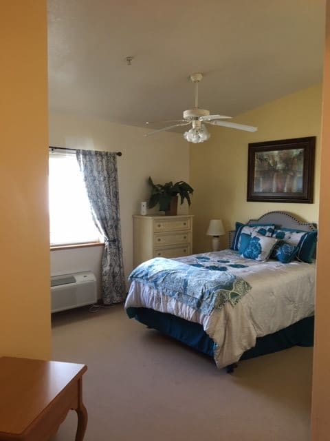 A cozy bedroom featuring a bed with blue and white bedding, a dresser, a window with curtains, and a ceiling fan.