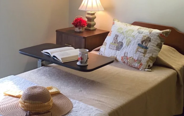 A cozy bedroom with a nightstand, lamp, book, and a mug on an over-bed table.