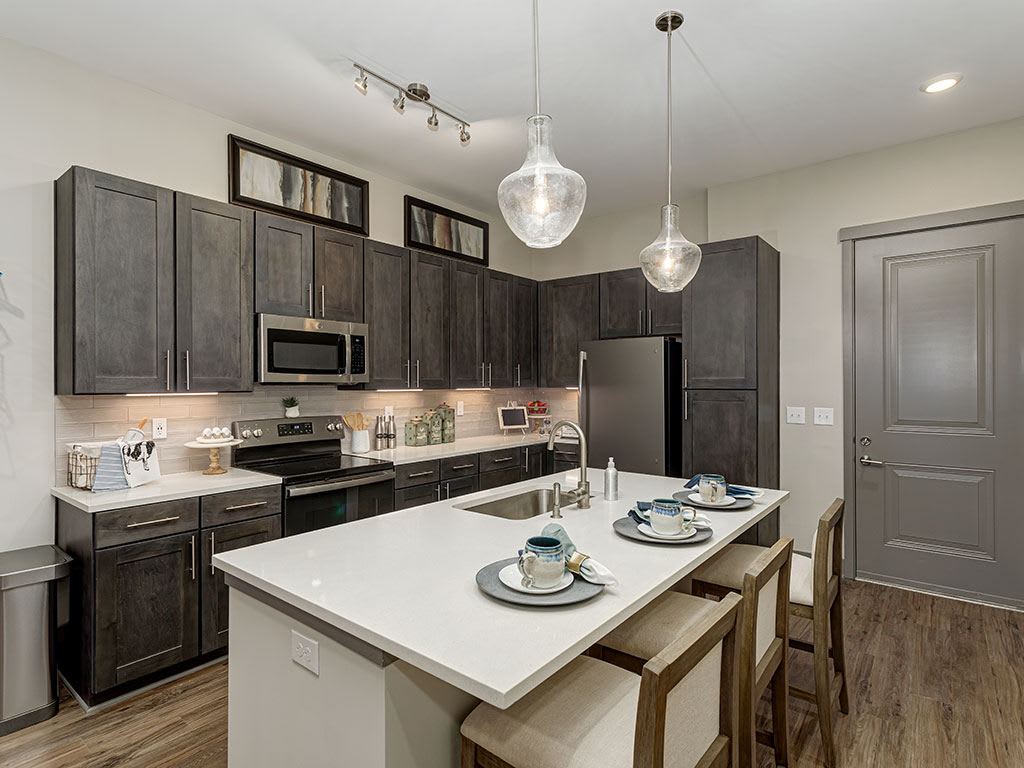 A modern kitchen featuring a central island with bar stools, pendant lighting, stainless steel appliances, and dark cabinetry.