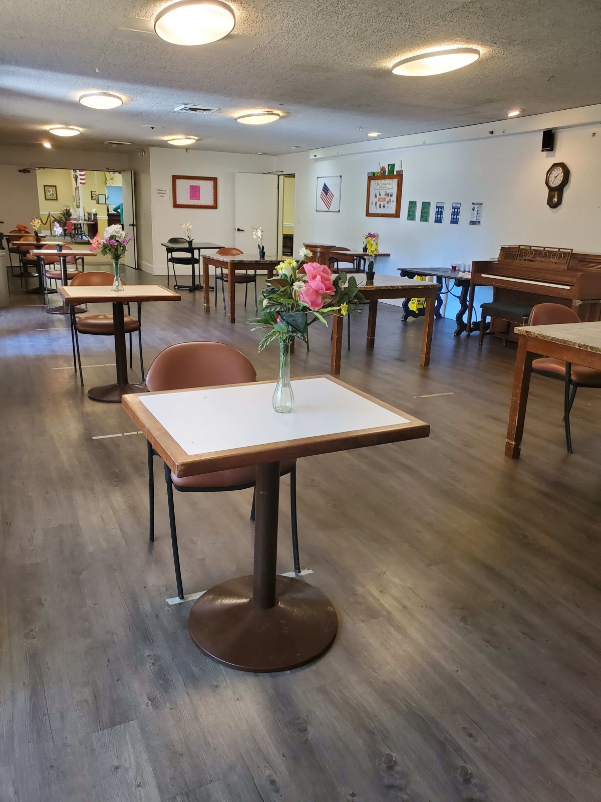 A neatly arranged dining room with tables, chairs, and a vase of flowers, featuring a piano in the corner.