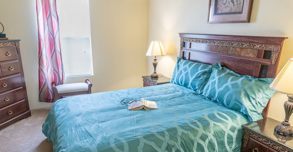 A cozy bedroom featuring a neatly made bed with teal bedding, a bedside lamp, and an open book.