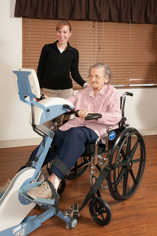 An elderly person in a wheelchair using an exercise machine with the assistance of a caregiver.