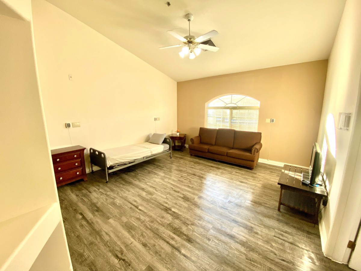 A spacious living room with wooden flooring, featuring a sofa, bed, window, and ceiling fan.