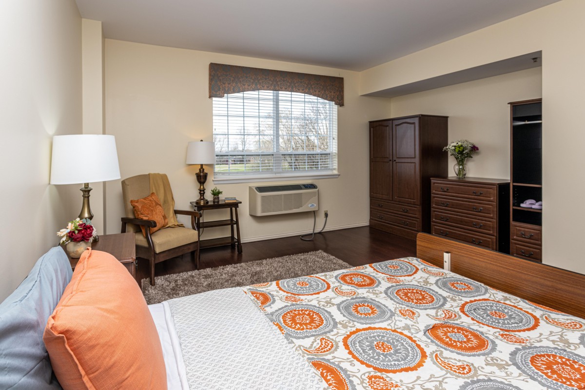A cozy bedroom with a patterned bedspread, wooden furniture, and a large window allowing natural light.