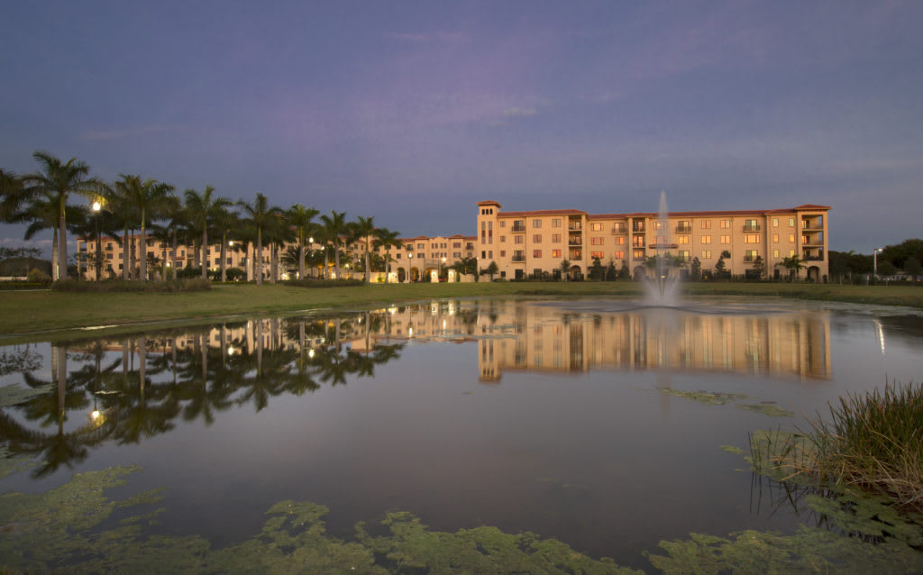 A tranquil sunset view of a building reflected in a lake, surrounded by palm trees.