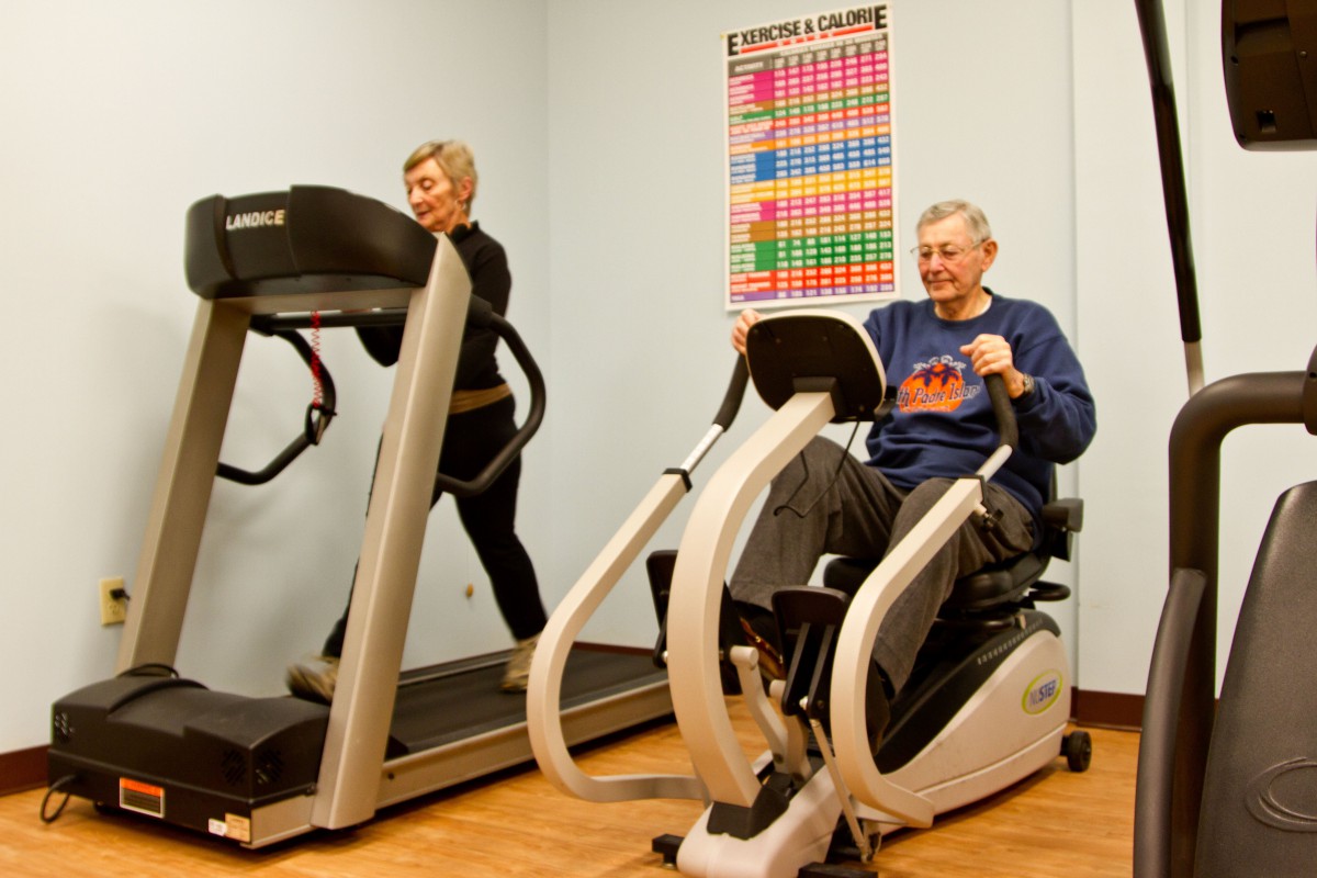 Two elderly individuals exercising on gym equipment, with one using a treadmill and the other on a stationary bike.
