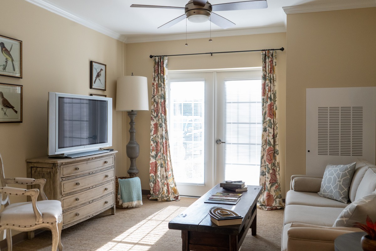 A cozy living room with a television, sofa, and coffee table, illuminated by natural light through large windows.