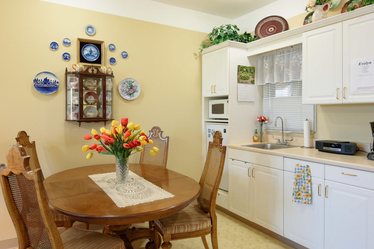 A cozy kitchen with a dining area, featuring a wooden table and chairs, a vase of flowers, and decorative wall elements.
