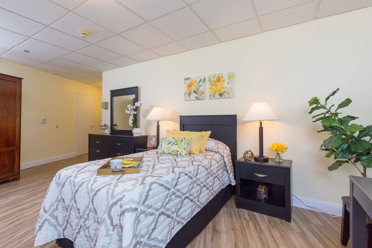 A well-decorated bedroom with a single bed, featuring a patterned comforter, pillows, and matching nightstands with lamps. The room has wall art above the bed and a dresser with a mirror.