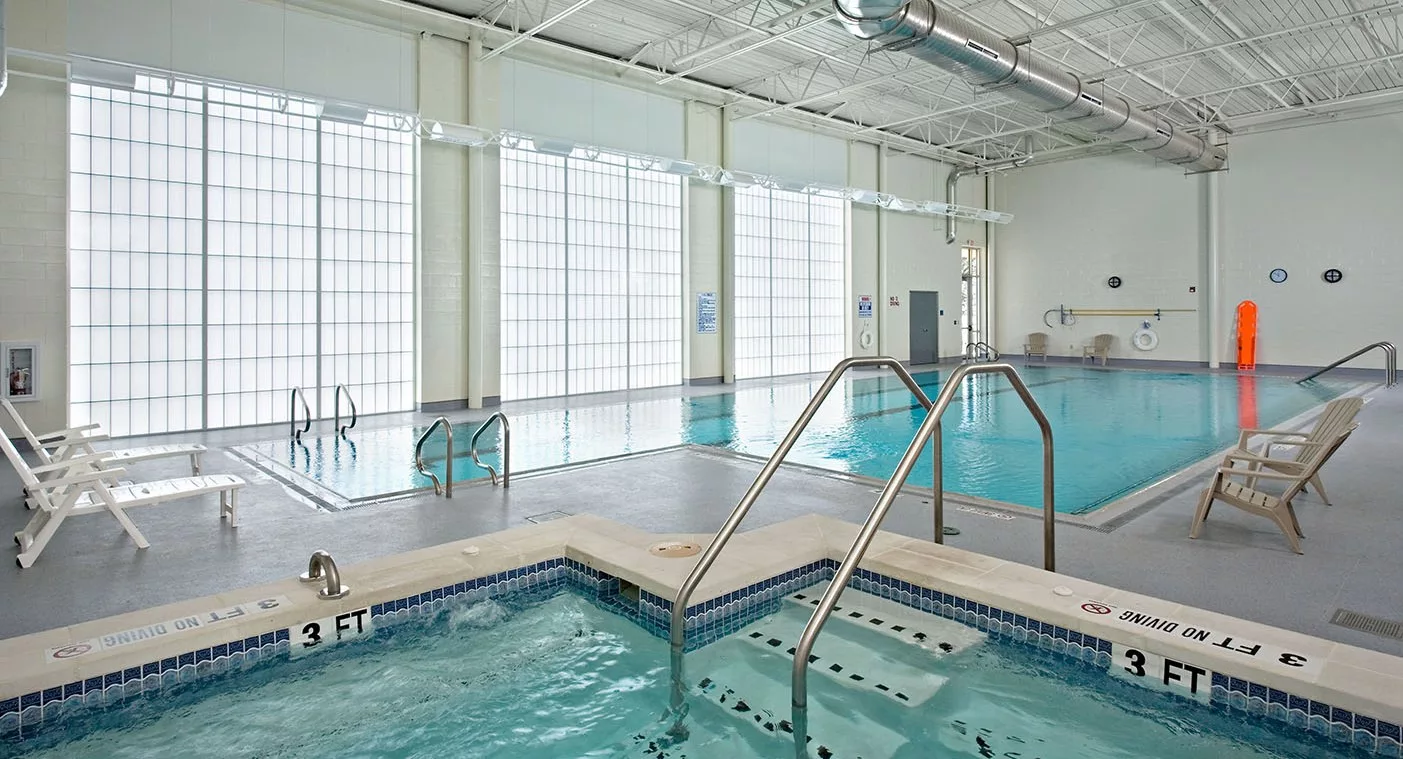 An indoor swimming area with a large pool and adjacent jacuzzi, surrounded by tall windows.