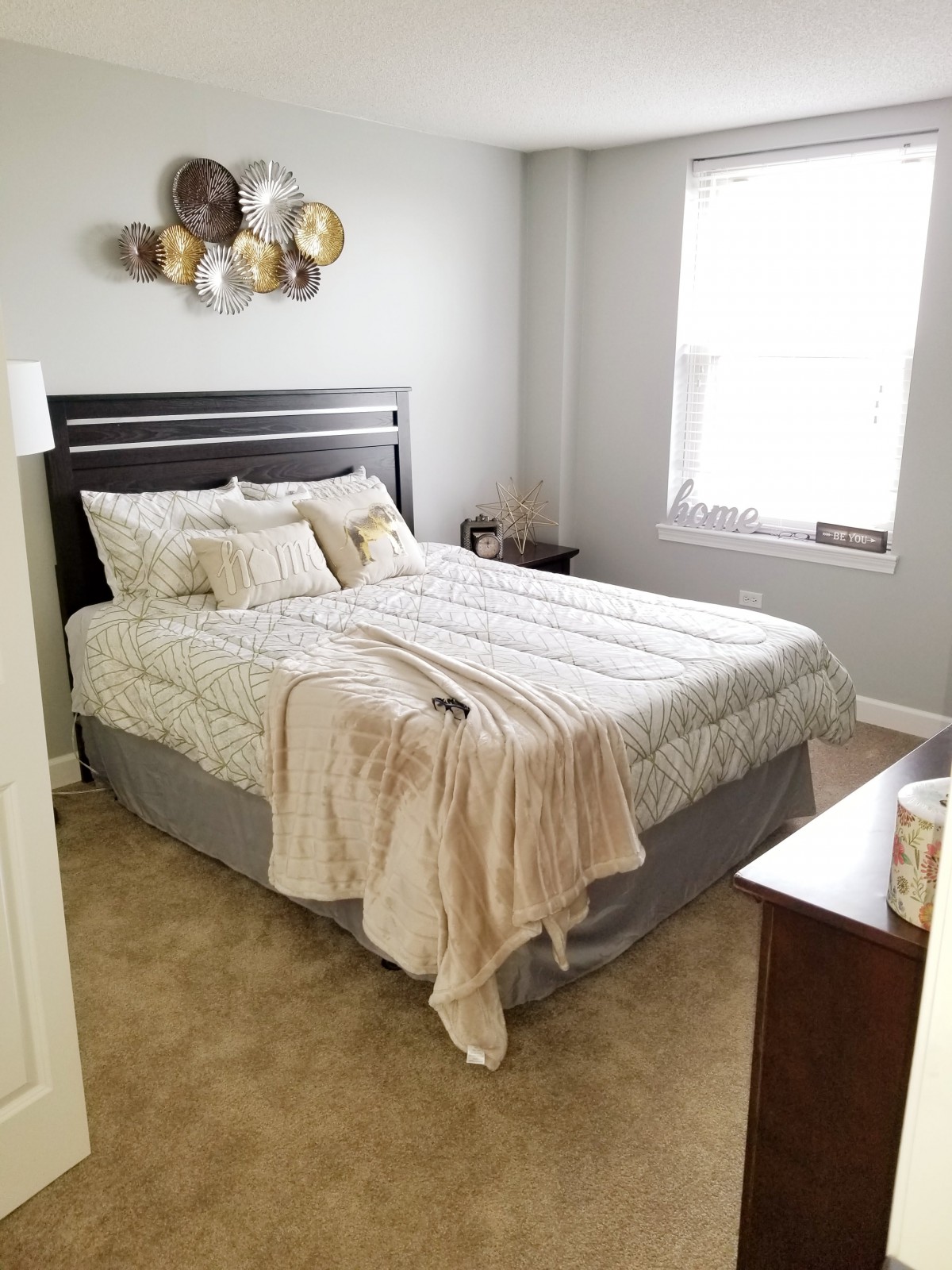 A cozy bedroom with a neatly made bed, decorative pillows, and a beige blanket draped over it. The room is brightened by natural light from a window.