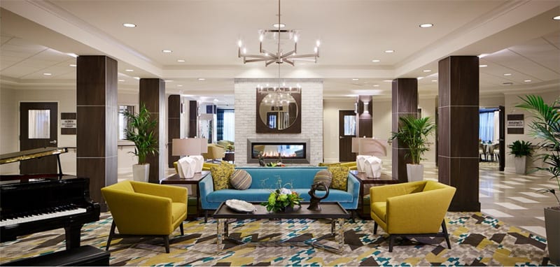 A modern living room featuring a blue sofa and yellow armchairs, centered around a glass coffee table and a stylish chandelier. The room includes decorative elements like lamps and a fireplace.
