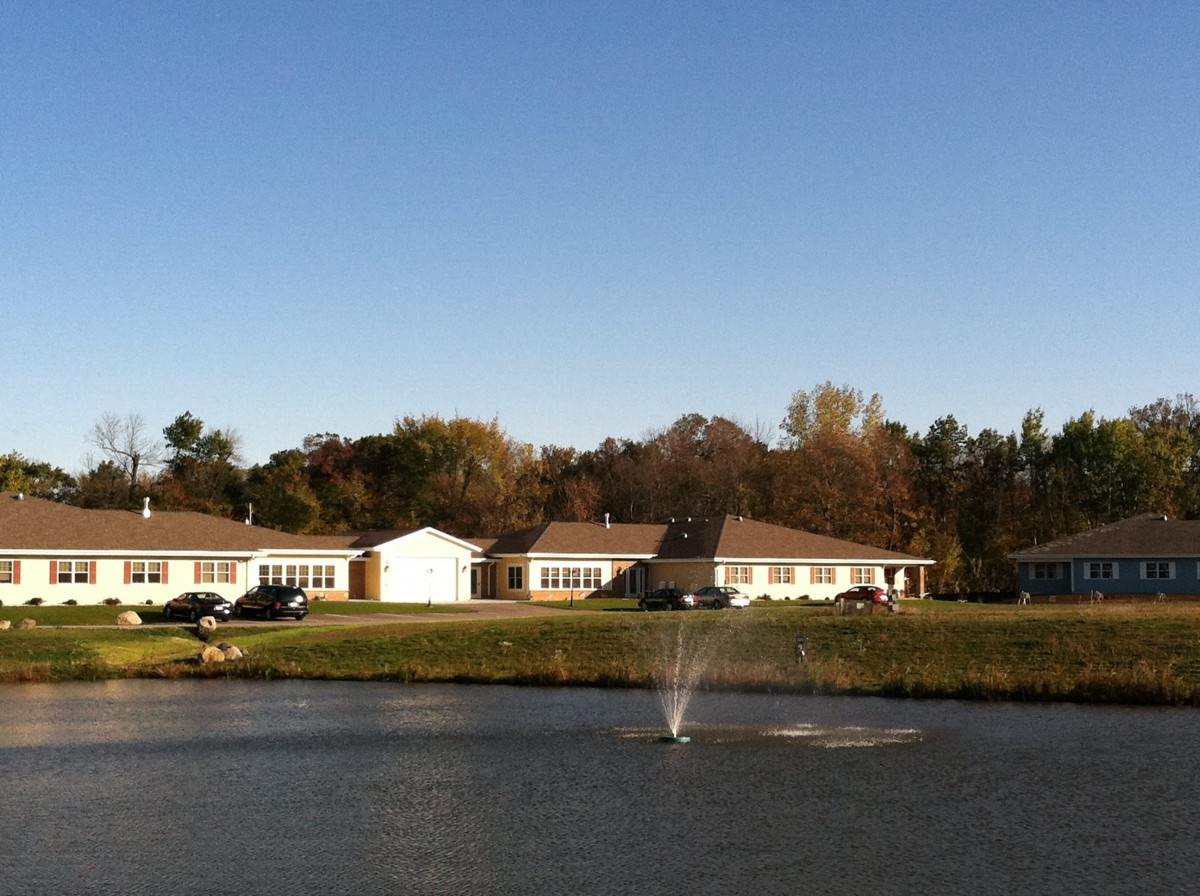A serene lakeside view with a fountain, surrounded by houses and trees under a clear blue sky.