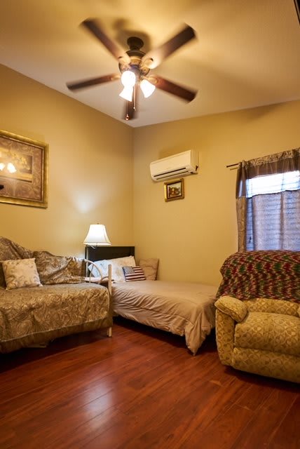 A cozy living room with a sofa, armchair, and a bed, featuring a ceiling fan and air conditioner.