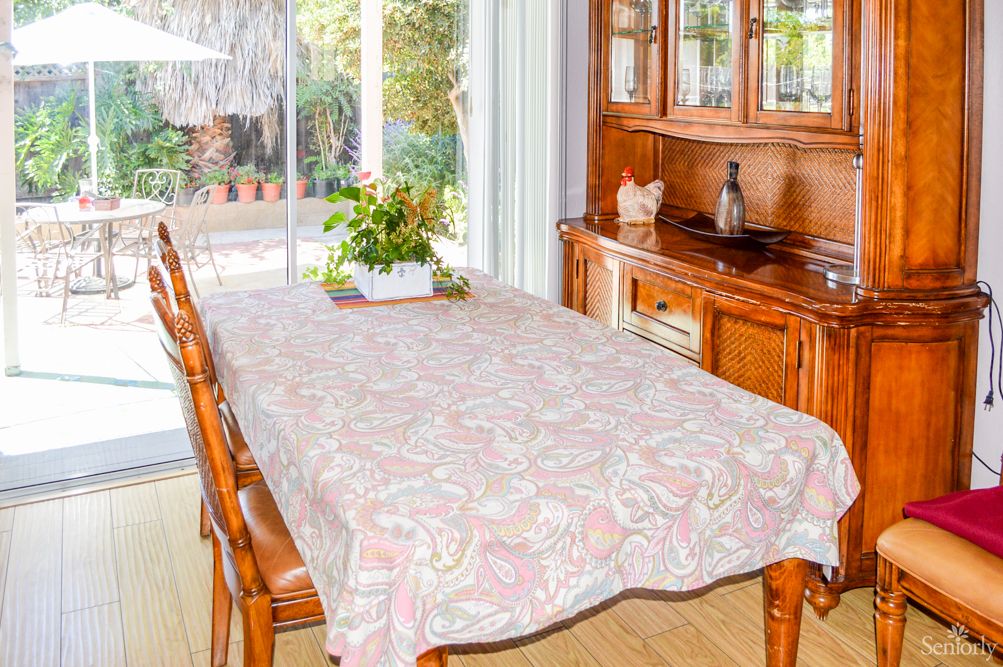 A cozy dining room with a table covered by a patterned tablecloth, surrounded by wooden chairs, and a cabinet in the background.