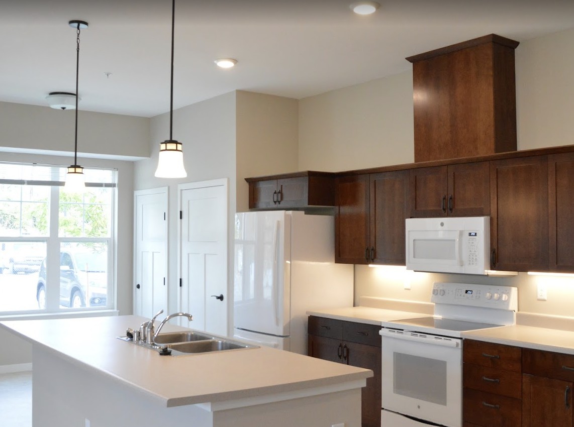 A modern kitchen featuring a central island counter and wooden cabinetry, equipped with white appliances and pendant lighting.