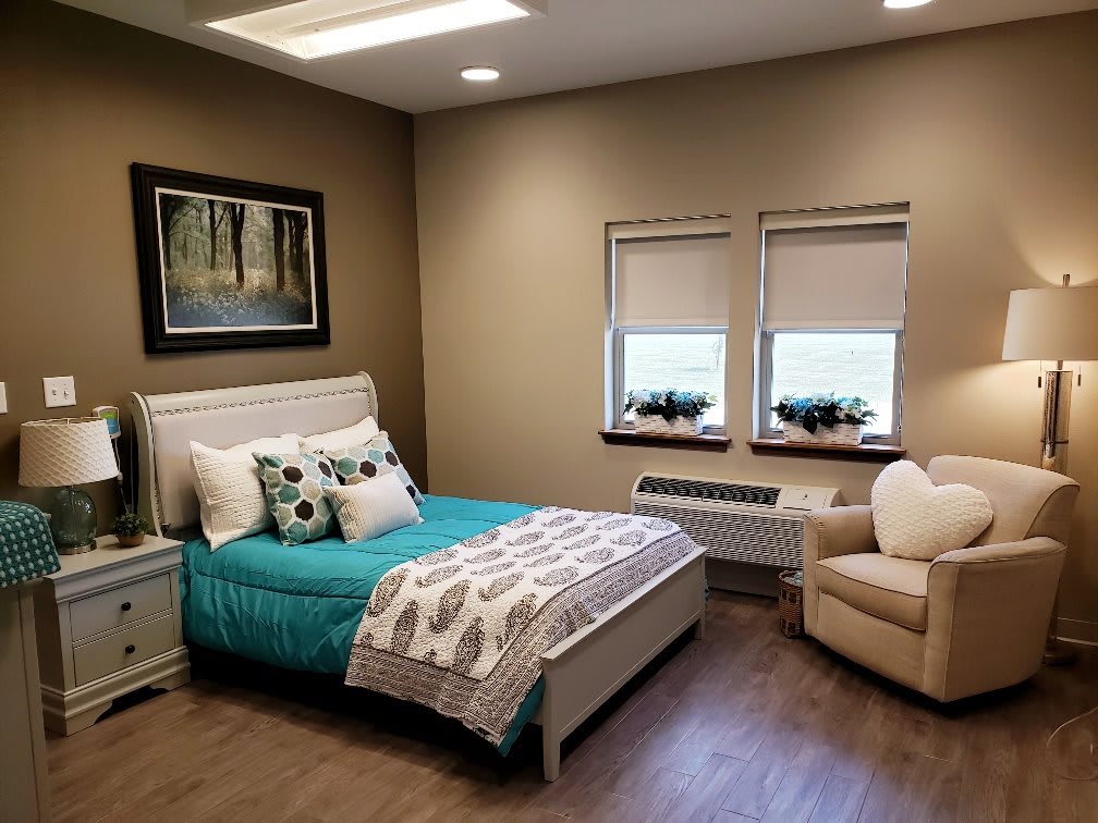 A cozy bedroom featuring a neatly made bed, an upholstered chair, and two windows adorned with blinds. The room is decorated with a nature-themed artwork.