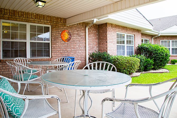 A cozy patio with a glass table and metal chairs, set against a brick wall and overlooking a small garden.