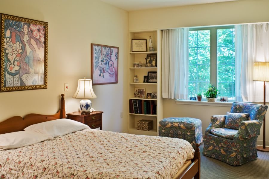 A cozy bedroom with a neatly made bed, a lamp on the nightstand, and a window with a view of greenery. The room features bookshelves filled with items and artwork on the walls.