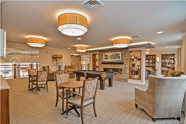A cozy lounge area with tables, chairs, and bookshelves under warm lighting.