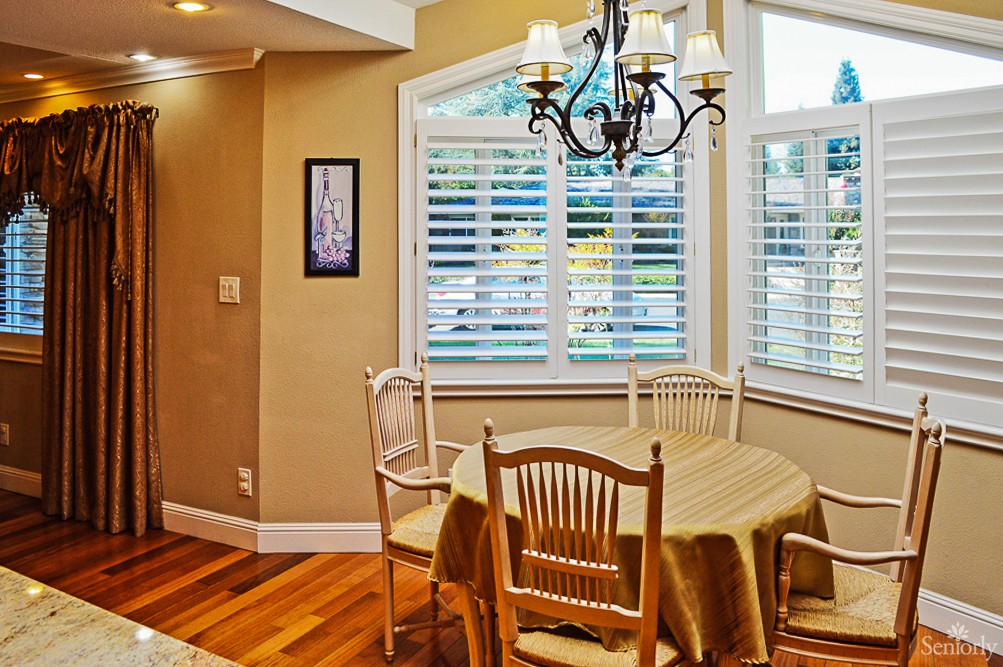 A cozy dining area with a round table, chairs, and a chandelier in front of large windows.