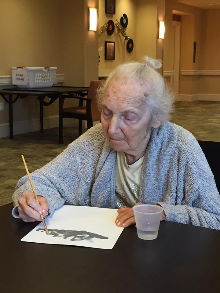 An elderly woman engages in painting, creating art with a brush indoors.