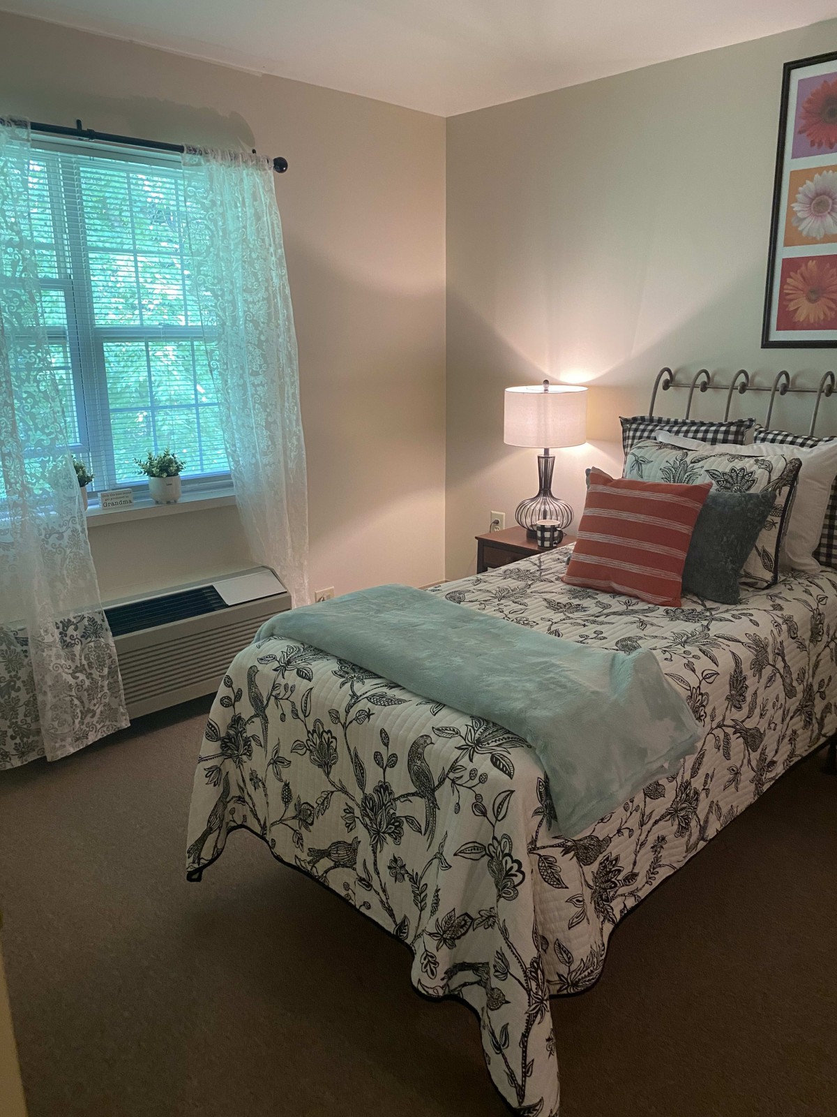 A cozy bedroom with a single bed, adorned with floral bedding and pillows, next to a bedside lamp and window with sheer curtains.