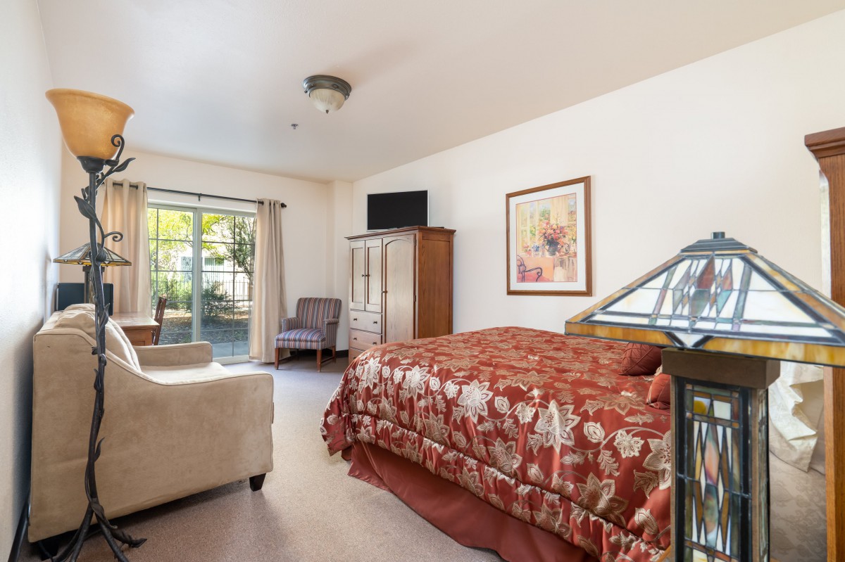 A cozy bedroom featuring a bed with a floral cover, a window with curtains, and various furniture pieces including a chair and dresser.