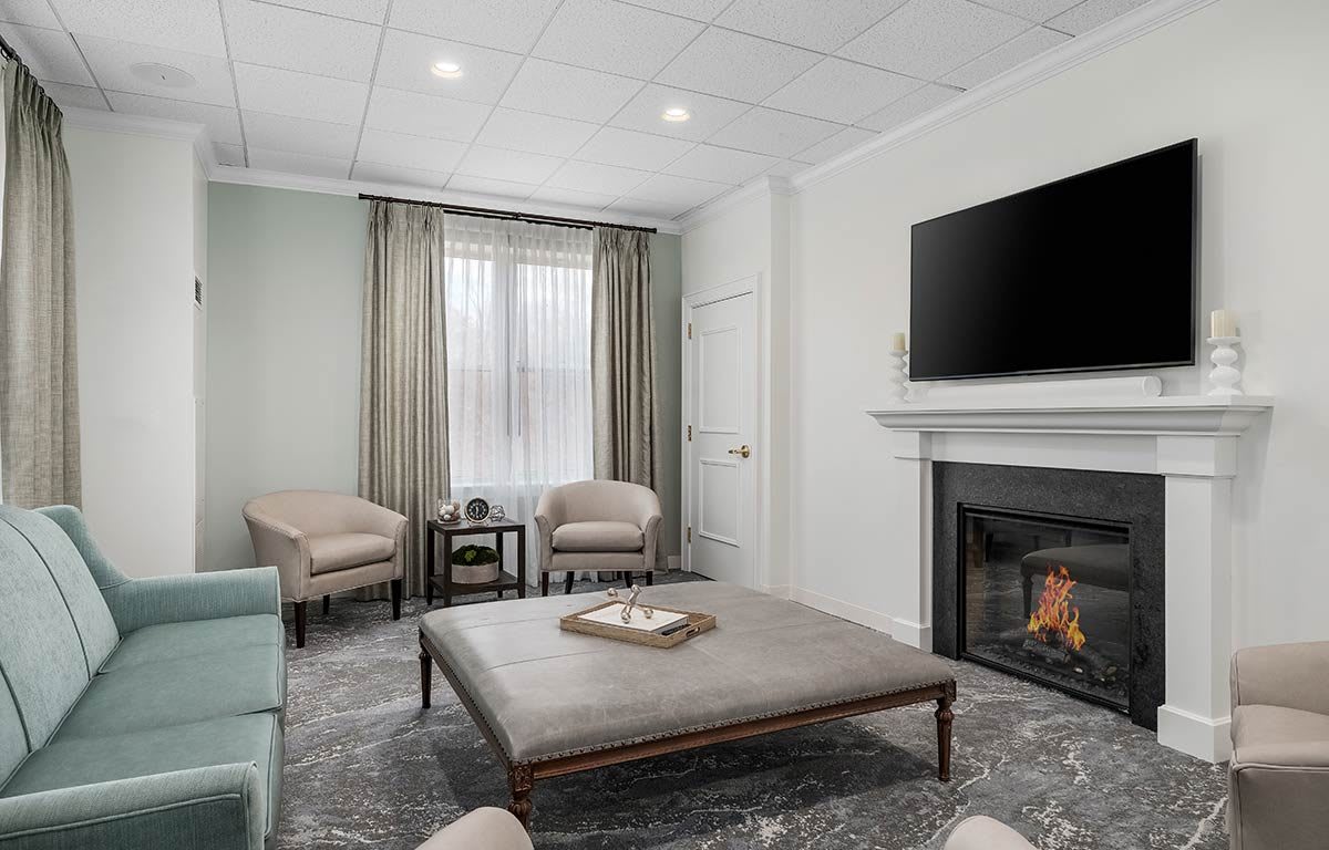 A modern living room featuring a large ottoman, cozy chairs, a fireplace, and a wall-mounted television.