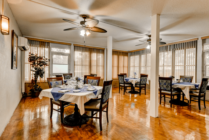 A well-furnished dining area with elegantly set tables and ceiling fans, featuring a wooden floor and large windows with curtains.