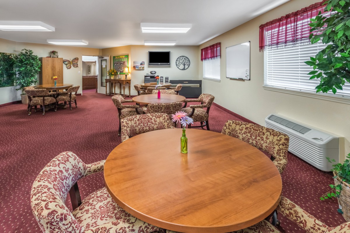 A cozy dining area with round wooden tables and patterned chairs, featuring carpeted floors and windows with red valances.