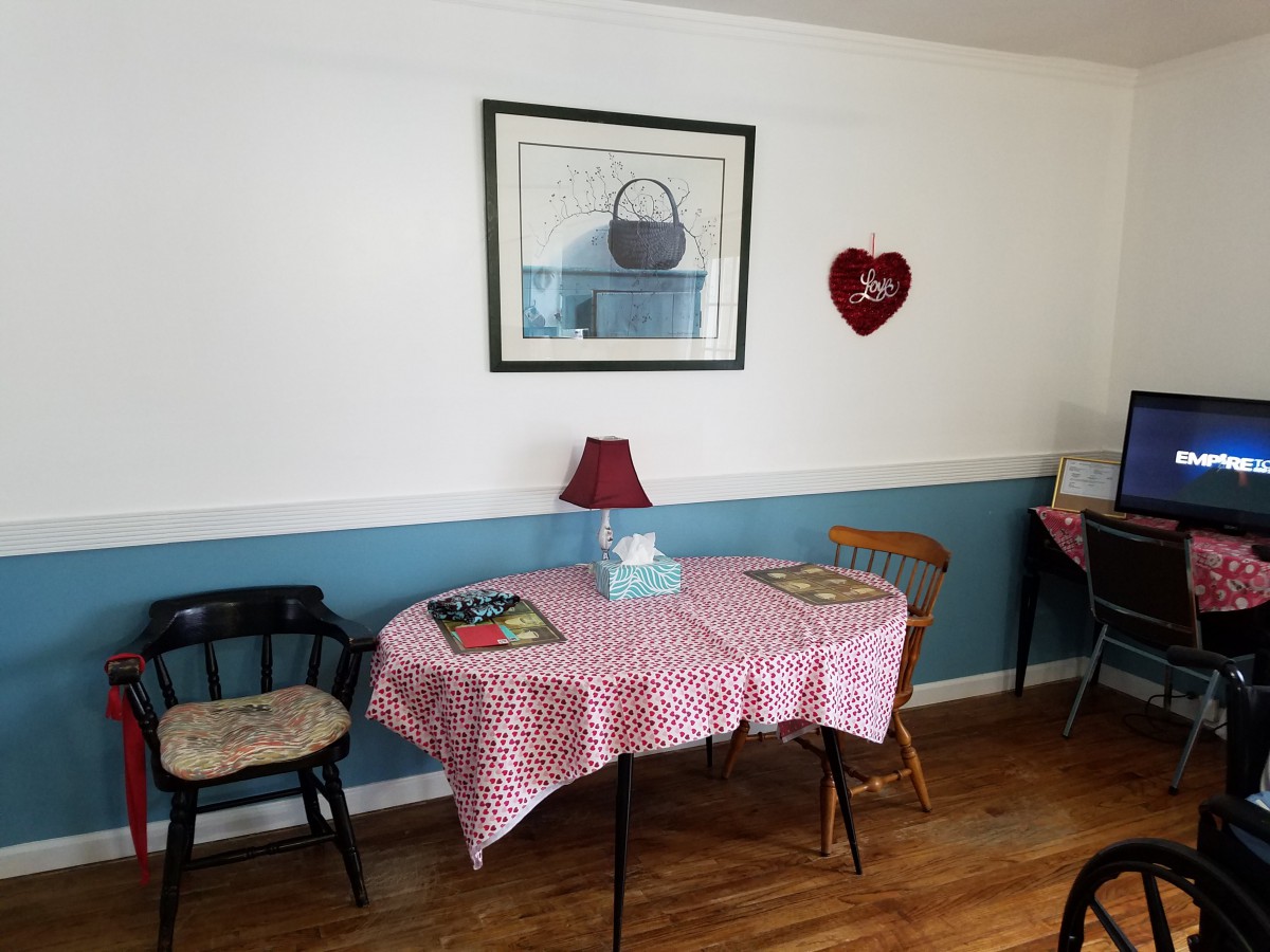 A cozy dining area with a table covered by a patterned tablecloth, surrounded by chairs and decorated with wall art and a small lamp.