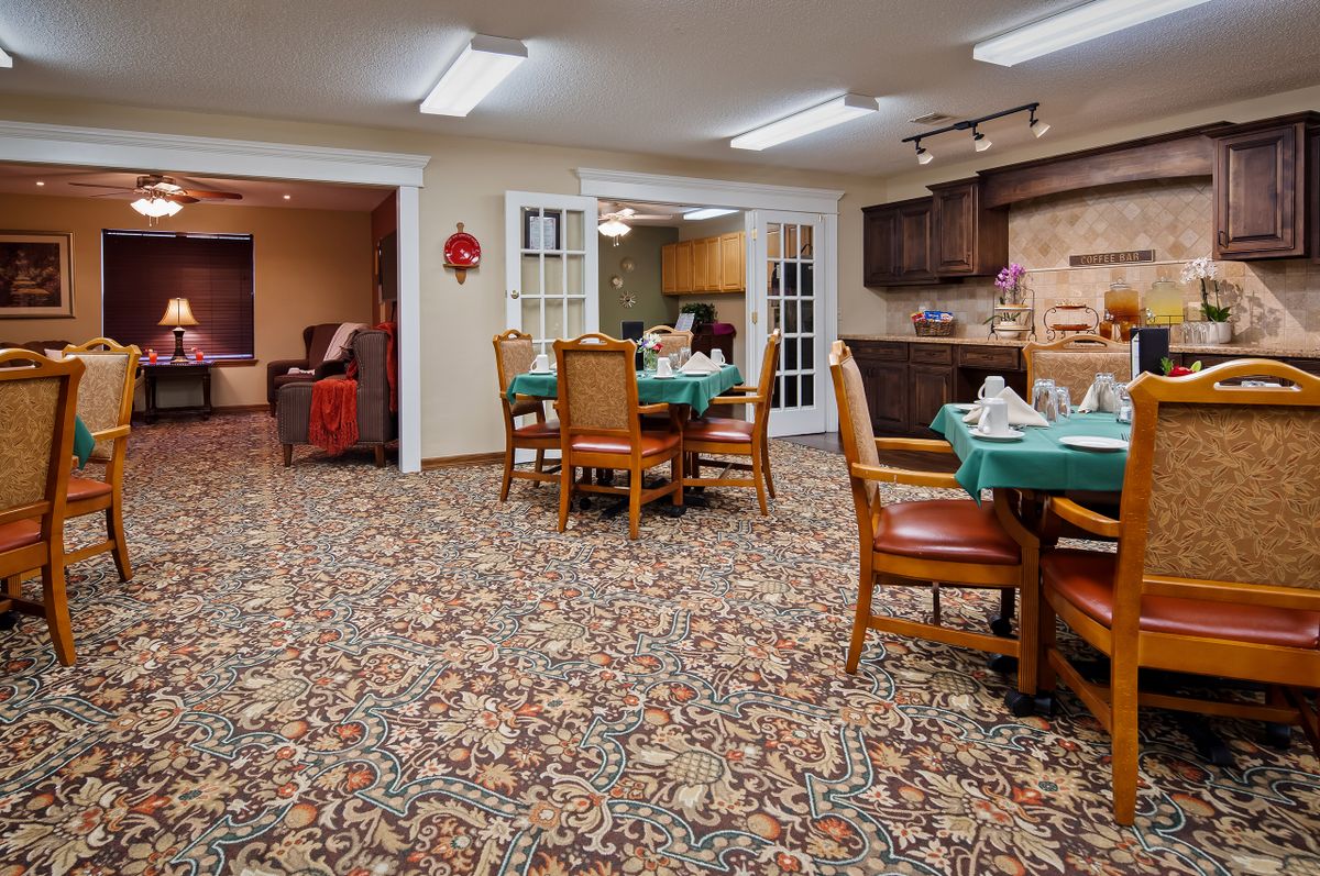 A cozy dining area with a carpeted floor, set with tables and chairs, leading to a kitchen space.