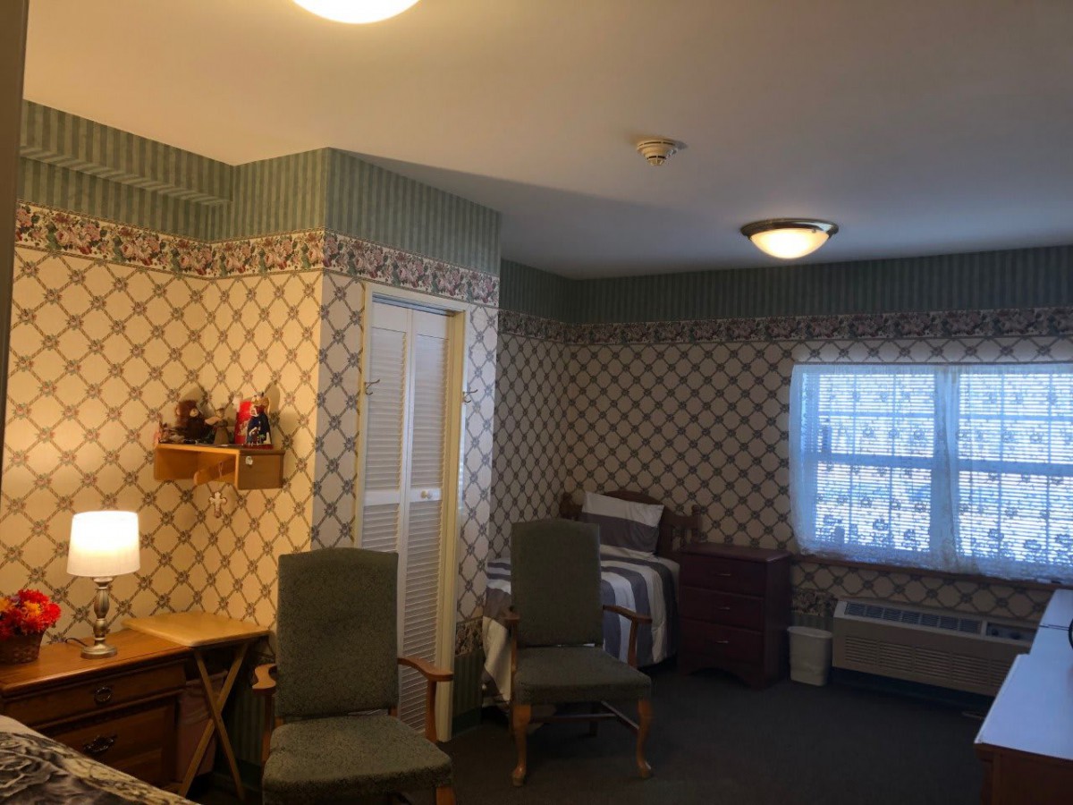 A room with patterned wallpaper, featuring chairs, a bed, and a large window.