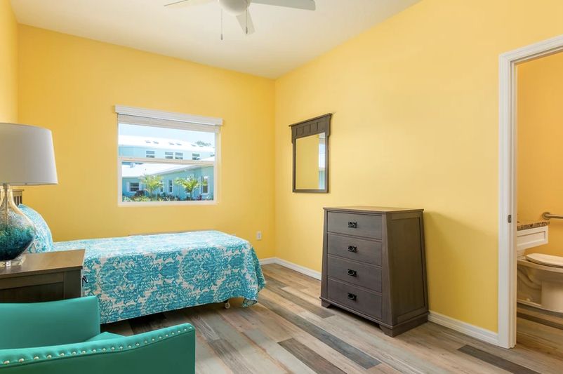 A cozy bedroom with bright yellow walls, featuring a single bed covered in a patterned turquoise bedspread. The room includes a dark wooden dresser and a mirror, with natural light streaming through the window onto the wooden flooring.