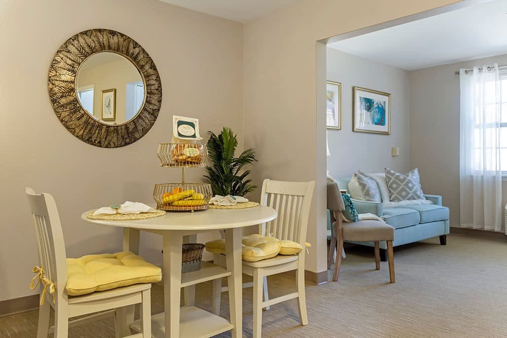 A cozy dining area with a round table and chairs, featuring a decorative mirror and an adjacent living room.