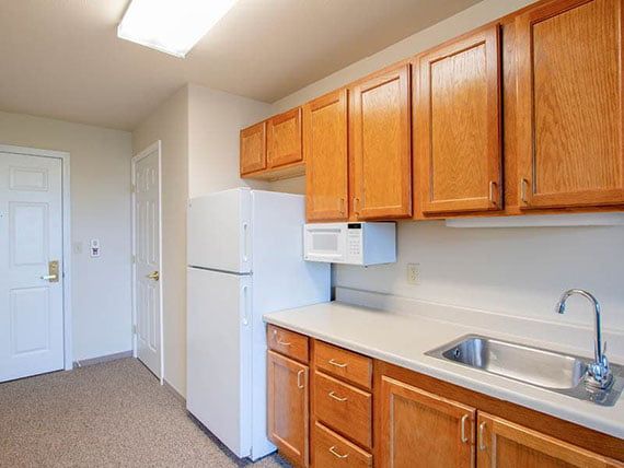 A simple kitchen with wooden cabinets, a refrigerator, microwave, and sink.