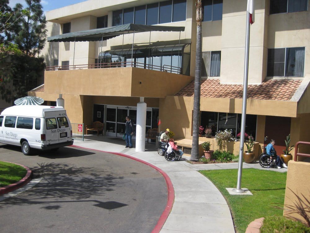 A hospital entrance with a van parked nearby and people gathered outside.