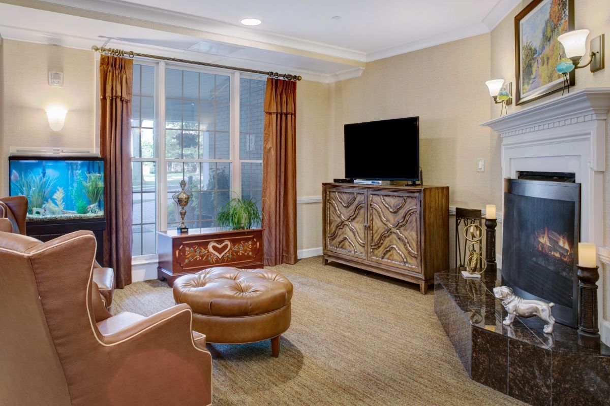 A cozy living room featuring a television on a decorative cabinet, an armchair with an ottoman, and large windows with curtains.
